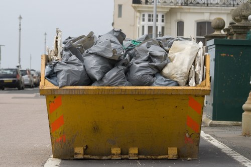 A large yellow industrial skip positioned on a paved street in Wanstead, filled with black and white heavy-duty rubbish bags made of thick plastic material. The bags are piled unevenly, some leaning against the edges of the skip, with a few slightly bulging indicating contents inside. The skip is situated near the edge of a residential or commercial area, with parked cars visible in the background and a row of classic-style buildings with bay windows and decorative facades on the opposite side of the street. The sky appears overcast, casting diffuse natural light over the scene, and the skip shows signs of dirt and grime from previous use, consistent with waste removal services by Commercial Waste Removal Wanstead. The street setting suggests a typical suburban or urban environment where rubbish collection or clearance services are routinely conducted.