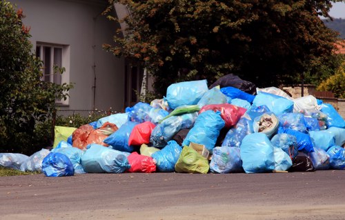 A large accumulation of assorted rubbish bags, predominantly in shades of blue, with some red, green, orange, and white, piled on the side of a paved road in front of a white building. The bags appear to contain household waste and are stacked haphazardly, spilling onto the pavement and partially obscuring the building's exterior. Behind the pile, there are green bushes and a tree with dark leaves, indicating a residential or suburban area. The scene is outdoors, under natural light, with the bags likely awaiting collection or removal by waste management services such as those offered by Commercial Waste Removal Wanstead, serving the local area around Wanstead and nearby postcode districts. The background includes a portion of a window with white framing and a partial view of a garden or yard area, contributing to the typical sight of unmanaged rubbish accumulation outside residential properties in the region.